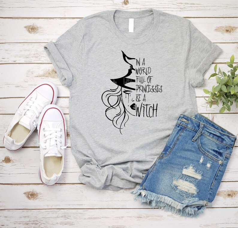 Peut inclure: Un t-shirt gris avec un graphique noir d'une sorci&egrave;re avec le texte "Dans un monde plein de princesses, sois une sorci&egrave;re".