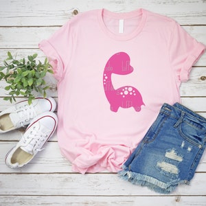 以下が含まれることがあります： ピンクのTシャツに、ピンクの恐竜のシルエットのグラフィックがプリントされています。恐竜には白い水玉模様があり、シャツには白い文字で「Little Designs」とプリントされています。