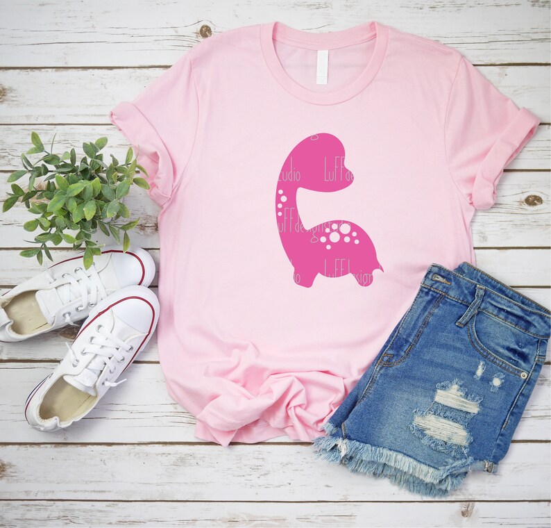 以下が含まれることがあります： ピンクのTシャツに、ピンクの恐竜のシルエットのグラフィックがプリントされています。恐竜には白い水玉模様があり、シャツには白い文字で「Little Designs」とプリントされています。