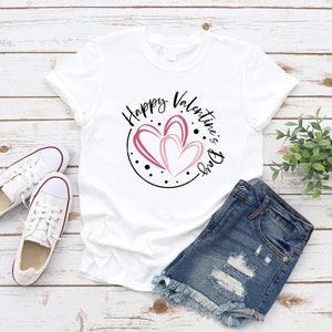 Puede incluir: Camiseta blanca con un dise&ntilde;o circular negro con el texto "Happy Valentine's Day" y tres corazones rosas. La camiseta est&aacute; colocada sobre una superficie de madera con un par de zapatillas blancas y unos pantalones cortos de mezclilla.