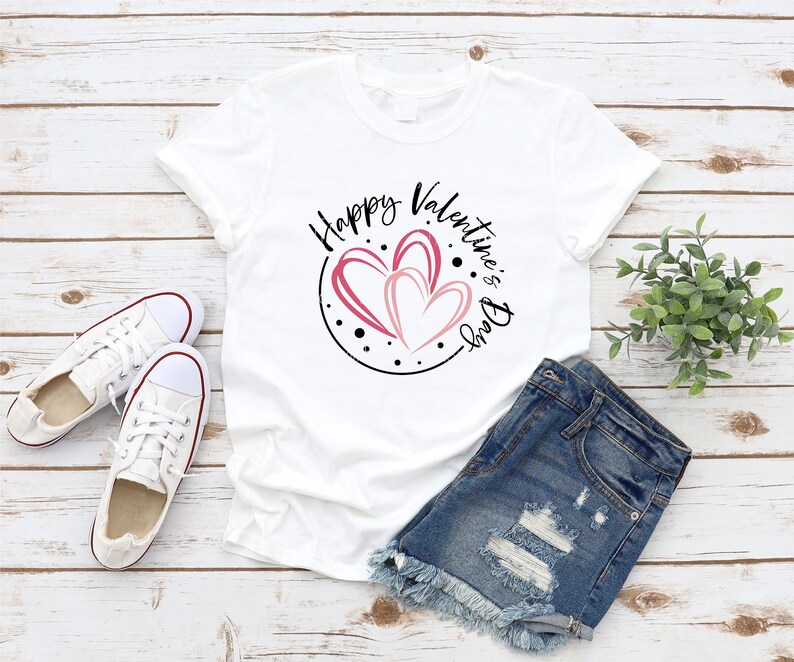 Puede incluir: Camiseta blanca con un dise&ntilde;o circular negro con el texto "Happy Valentine's Day" y tres corazones rosas. La camiseta est&aacute; colocada sobre una superficie de madera con un par de zapatillas blancas y unos pantalones cortos de mezclilla.