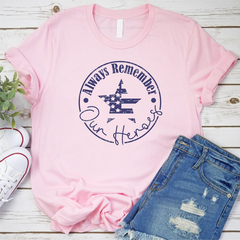 May include: Pink t-shirt with a blue distressed circle graphic featuring a star with an American flag design and the text "Always Remember Our Heroes".