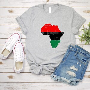 May include: A gray t-shirt with a red, black, and green outline of the continent of Africa. The shirt is laid flat on a wooden surface with a pair of white sneakers and a pair of blue denim shorts.