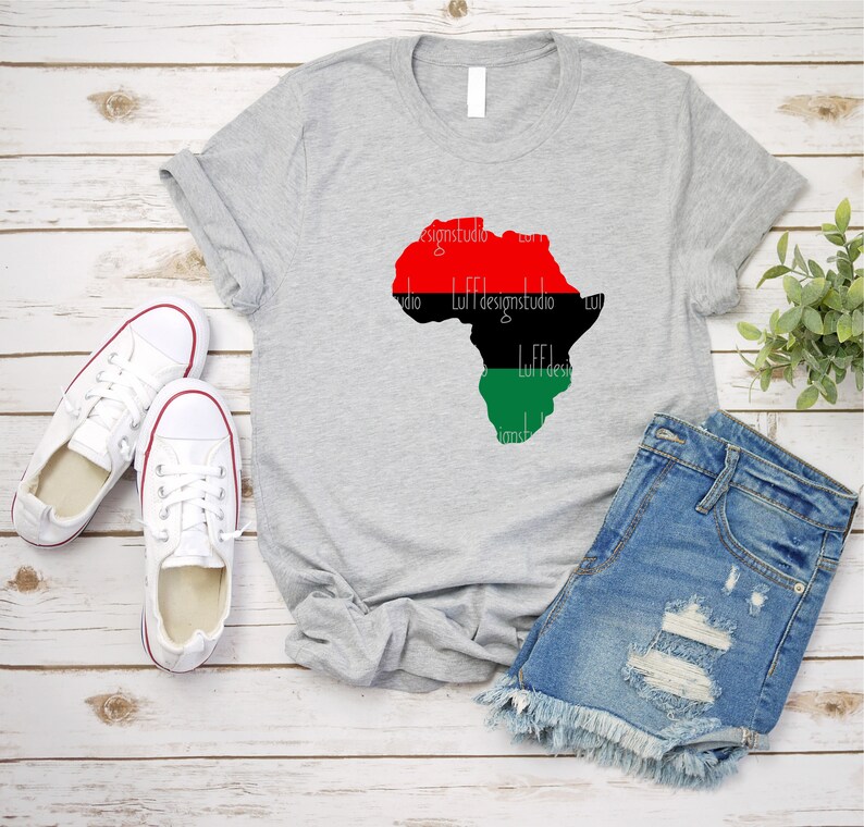 May include: A gray t-shirt with a red, black, and green outline of the continent of Africa. The shirt is laid flat on a wooden surface with a pair of white sneakers and a pair of blue denim shorts.