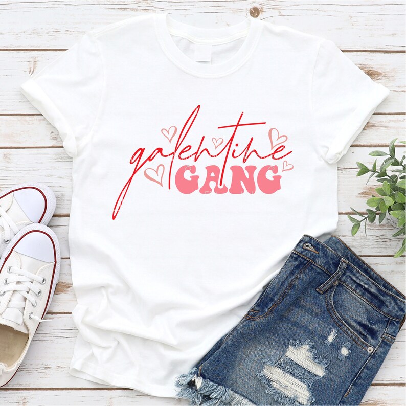 May include: White t-shirt with red and pink lettering that says "galentine gang" with pink hearts around the text.
