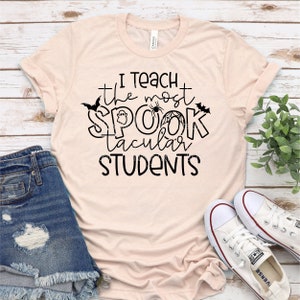 以下が含まれることがあります： 薄いピンクのTシャツに、黒い文字で「私は最も素晴らしい生徒を教えます」と書かれています。テキストは、幽霊、クモ、コウモリで飾られています。