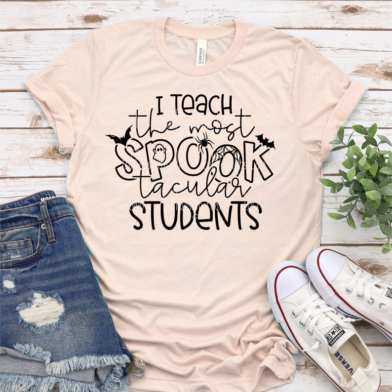 以下が含まれることがあります： 薄いピンクのTシャツに、黒い文字で「私は最も素晴らしい生徒を教えます」と書かれています。テキストは、幽霊、クモ、コウモリで飾られています。