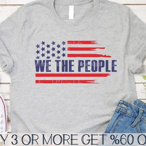 Op de afbeelding: Een grijs t-shirt met een verweerd Amerikaans vlagontwerp en de tekst "WE THE PEOPLE" in marineblauw. Het vlagontwerp bevat rode strepen en blauwe sterren. Het shirt wordt getoond met een spijkerbroek.