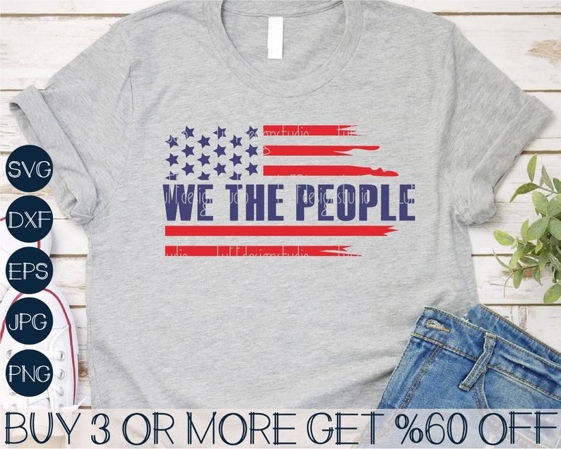 Op de afbeelding: Een grijs t-shirt met een verweerd Amerikaans vlagontwerp en de tekst "WE THE PEOPLE" in marineblauw. Het vlagontwerp bevat rode strepen en blauwe sterren. Het shirt wordt getoond met een spijkerbroek.