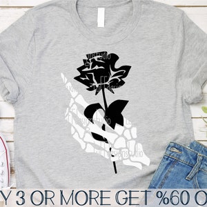 May include: A black and white graphic design of a skeleton hand holding a black rose. The design is on a gray t-shirt.