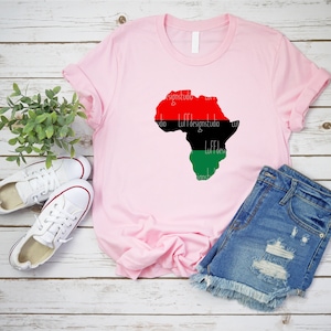 May include: A pink t-shirt with a red, black, and green outline of the continent of Africa. The shirt is laying on a wooden surface with a pair of white sneakers and blue denim shorts.