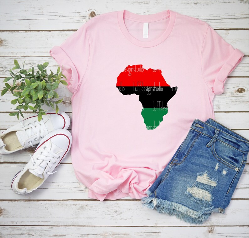 May include: A pink t-shirt with a red, black, and green outline of the continent of Africa. The shirt is laying on a wooden surface with a pair of white sneakers and blue denim shorts.