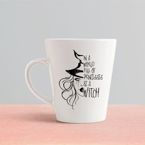 Peut inclure: Une tasse en c&eacute;ramique blanche avec une illustration en noir et blanc d'une sorci&egrave;re avec le texte "Dans un monde plein de princesses... sois une sorci&egrave;re".