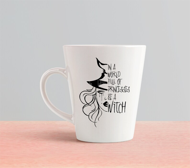 Peut inclure: Une tasse en c&eacute;ramique blanche avec une illustration en noir et blanc d'une sorci&egrave;re avec le texte "Dans un monde plein de princesses... sois une sorci&egrave;re".