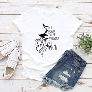 Peut inclure: T-shirt blanc avec un graphique noir d'une sorci&egrave;re avec le texte "Dans un monde plein de princesses, sois une sorci&egrave;re."