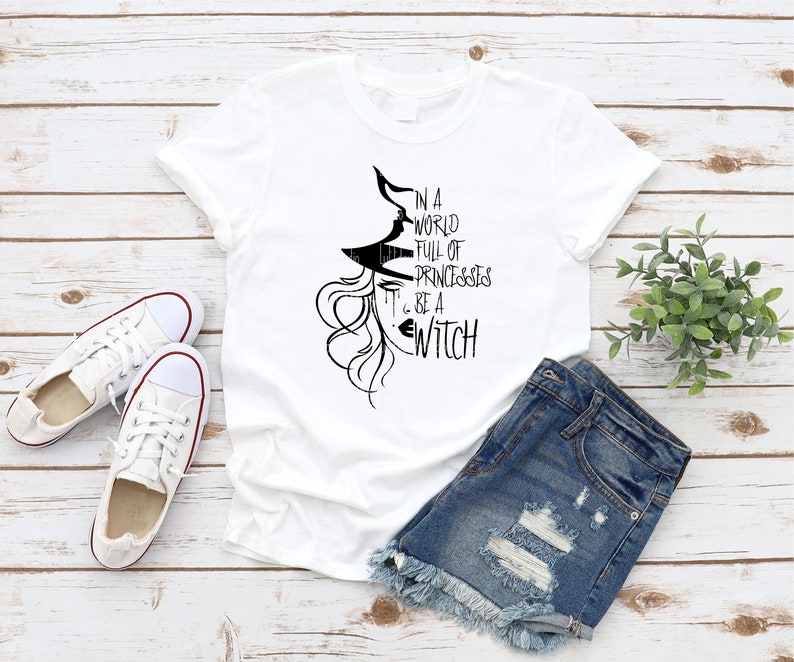 Peut inclure: T-shirt blanc avec un graphique noir d'une sorci&egrave;re avec le texte "Dans un monde plein de princesses, sois une sorci&egrave;re."