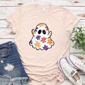 May include: A light pink t-shirt with a white ghost design. The ghost is covered in orange, purple, and yellow flowers.