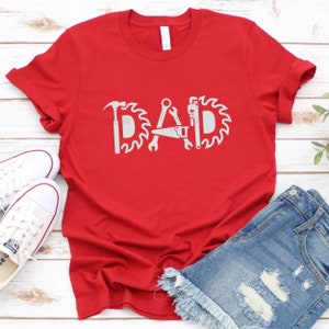 May include: Red t-shirt featuring a white graphic of the word "DAD" formed from tools such as a hammer, saw, wrench, and a circular saw blade.