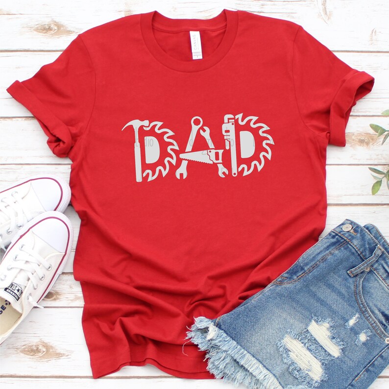May include: Red t-shirt featuring a white graphic of the word "DAD" formed from tools such as a hammer, saw, wrench, and a circular saw blade.