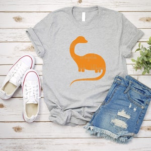 以下が含まれることがあります： オレンジ色の恐竜のシルエットデザインが施されたグレーのTシャツ。Tシャツは、白いスニーカーとデニムのショートパンツと一緒に木製の表面に置かれています。
