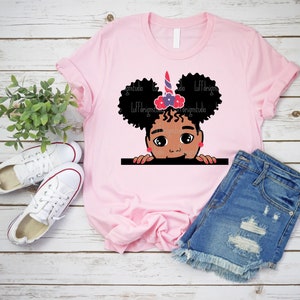 May include: Pink t-shirt featuring a graphic of a Black girl with curly hair peeking over a surface. She is wearing a unicorn headband with red, white and blue accents and a pink bow.