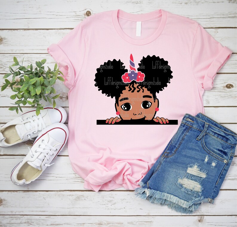 May include: Pink t-shirt featuring a graphic of a Black girl with curly hair peeking over a surface. She is wearing a unicorn headband with red, white and blue accents and a pink bow.