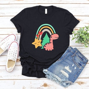 May include: Black t-shirt with a rainbow, a star, a tree, and a dinosaur. The dinosaur is pink and has a green tree behind it. The star is yellow and has a smiling face. The rainbow is multi-colored and has red hearts on it.