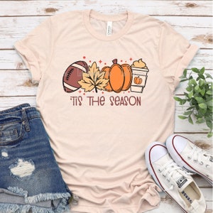 May include: A light pink t-shirt with a graphic design of a football, a pumpkin, a fall leaf, and a cup of coffee. The text "It's the season" is printed below the graphic.