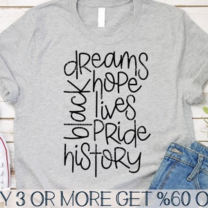 May include: A gray t-shirt with black text that reads "dreams hope black lives pride history". The text is in a handwritten style. The t-shirt is on a wooden surface with a pair of blue jeans and a pair of white sneakers.