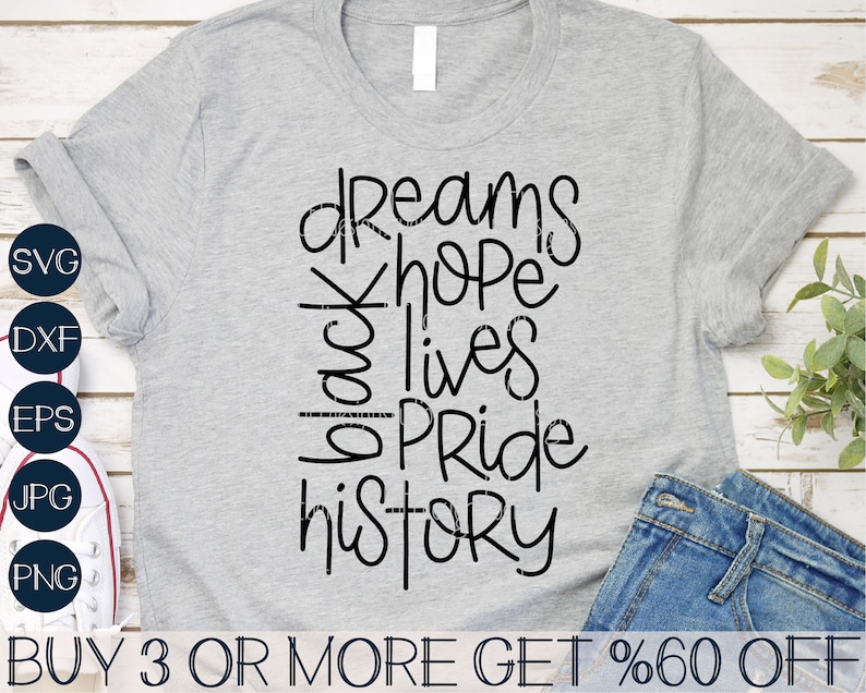 May include: A gray t-shirt with black text that reads "dreams hope black lives pride history". The text is in a handwritten style. The t-shirt is on a wooden surface with a pair of blue jeans and a pair of white sneakers.