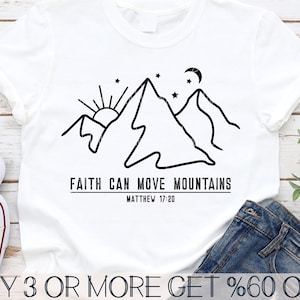Puede incluir: Una camiseta blanca con un dibujo de línea negra de montañas, un sol, una luna y estrellas. El texto "FAITH CAN MOVE MOUNTAINS" y "MATTHEW 17:20" está impreso debajo de la imagen.