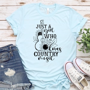 Puede incluir: Camiseta azul claro con el texto "Just a girl who loves country music" en negro. El dise&ntilde;o presenta una forma de guitarra con girasoles y hojas. La camiseta se combina con pantalones cortos de mezclilla y zapatillas de lona blancas.