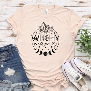 May include: A light pink t-shirt with a black graphic design of a circle with the text "Witchy mama" inside. The circle is surrounded by stars and crescent moons. There are crystals and flowers at the top of the circle.