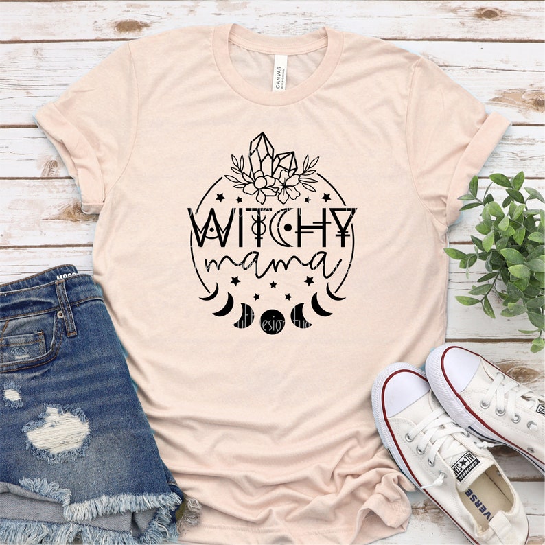 May include: A light pink t-shirt with a black graphic design of a circle with the text "Witchy mama" inside. The circle is surrounded by stars and crescent moons. There are crystals and flowers at the top of the circle.