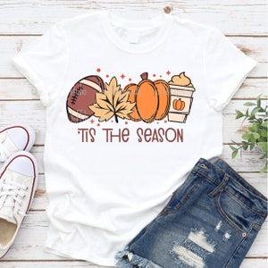 May include: White t-shirt with a graphic design of a football, fall leaves, pumpkins, and a coffee cup. The text "Tis the season" is printed below the graphic.