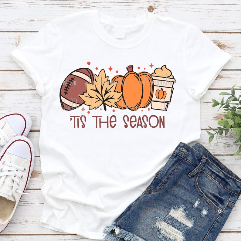 May include: White t-shirt with a graphic design of a football, fall leaves, pumpkins, and a coffee cup. The text "Tis the season" is printed below the graphic.