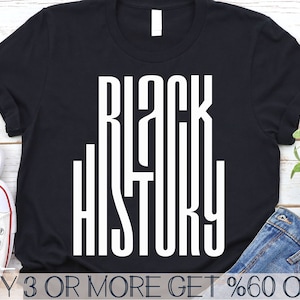 May include: Black t-shirt with white text that reads "Black History" in a bold, blocky font. The shirt is lying on a wooden surface with a pair of jeans and a pair of white trainers.