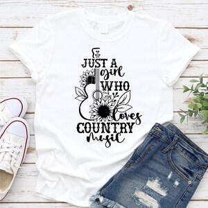 Puede incluir: Camiseta blanca con un gr&aacute;fico negro de una guitarra, girasoles y el texto "Just a girl who loves country music". La camiseta se combina con pantalones cortos de mezclilla y zapatillas blancas.