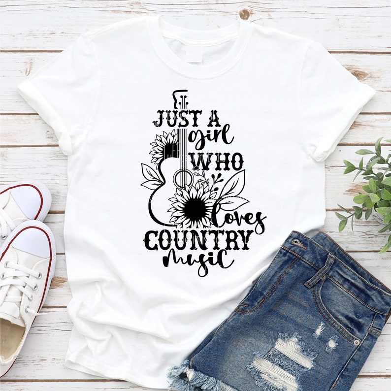 Puede incluir: Camiseta blanca con un gr&aacute;fico negro de una guitarra, girasoles y el texto "Just a girl who loves country music". La camiseta se combina con pantalones cortos de mezclilla y zapatillas blancas.