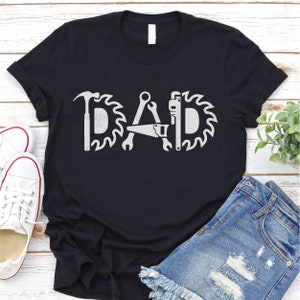 May include: Black t-shirt with a white graphic of the word "DAD" made up of tools like a hammer, saw, wrench, and a saw blade.