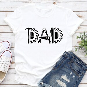 May include: White t-shirt with a black graphic design that says "DAD" with tools like a hammer, saw, wrench, and a pipe wrench incorporated into the letters.