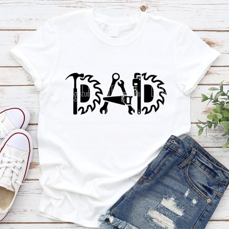 May include: White t-shirt with a black graphic design that says "DAD" with tools like a hammer, saw, wrench, and a pipe wrench incorporated into the letters.
