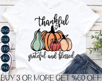 May include: A white t-shirt with the text "thankful grateful and blessed" in black script. There are three pumpkins in the design, one green and two orange. The pumpkins are stylized and have a vintage look.