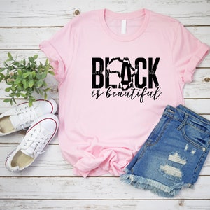 May include: Pink t-shirt with black text that reads "BLACK is beautiful". The text is styled with a silhouette of the continent of Africa.