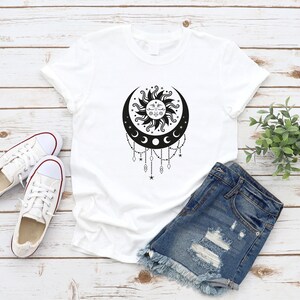 May include: White t-shirt with a black sun and moon design. The sun has a face and is surrounded by rays. The moon is crescent shaped and has three smaller moons hanging from it. There are stars and beads hanging from the bottom of the design.