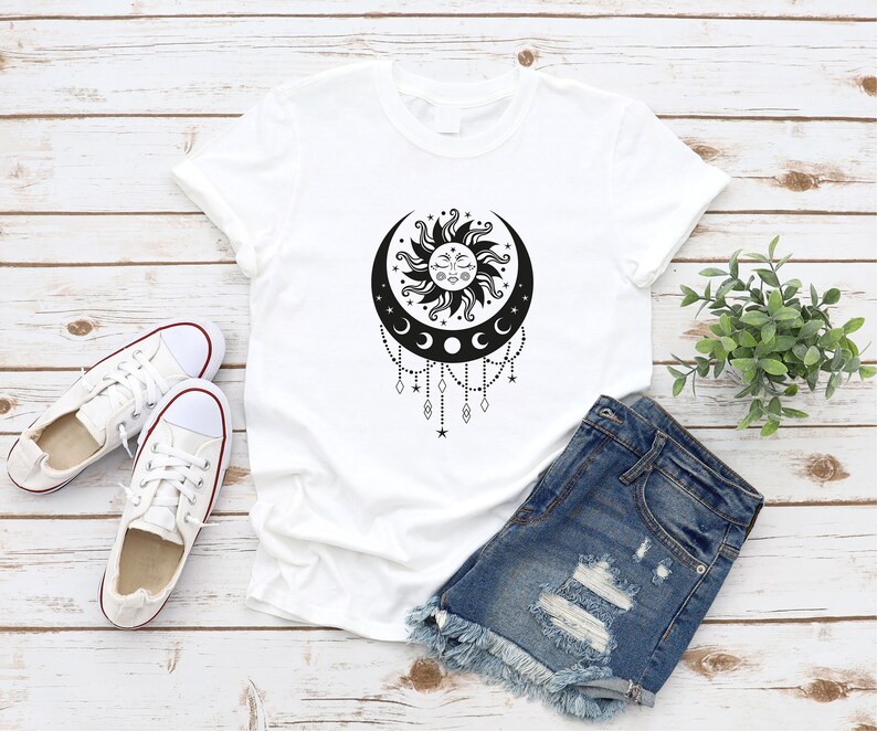 May include: White t-shirt with a black sun and moon design. The sun has a face and is surrounded by rays. The moon is crescent shaped and has three smaller moons hanging from it. There are stars and beads hanging from the bottom of the design.