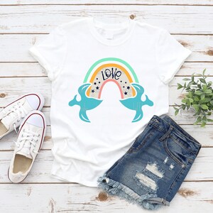 May include: A white t-shirt with a rainbow design and the word "Love" in a blue font. The rainbow has a yellow, orange, red, and blue color scheme. The t-shirt is laid out on a wooden surface with a pair of white sneakers and denim shorts.