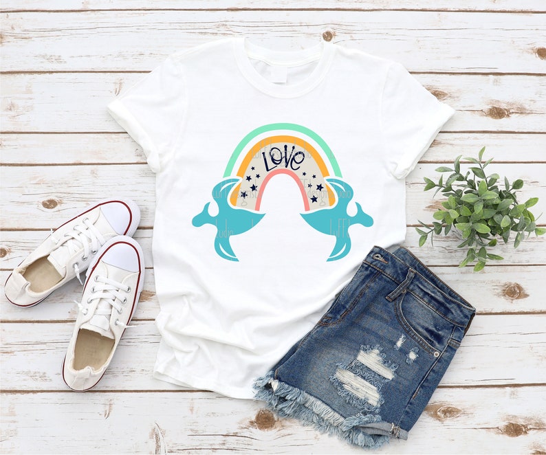 May include: A white t-shirt with a rainbow design and the word "Love" in a blue font. The rainbow has a yellow, orange, red, and blue color scheme. The t-shirt is laid out on a wooden surface with a pair of white sneakers and denim shorts.