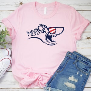 May include: Pink t-shirt with a shark graphic wearing sunglasses. The shark is made up of red, white, and blue stars and the text "Merica" is printed on the shirt.