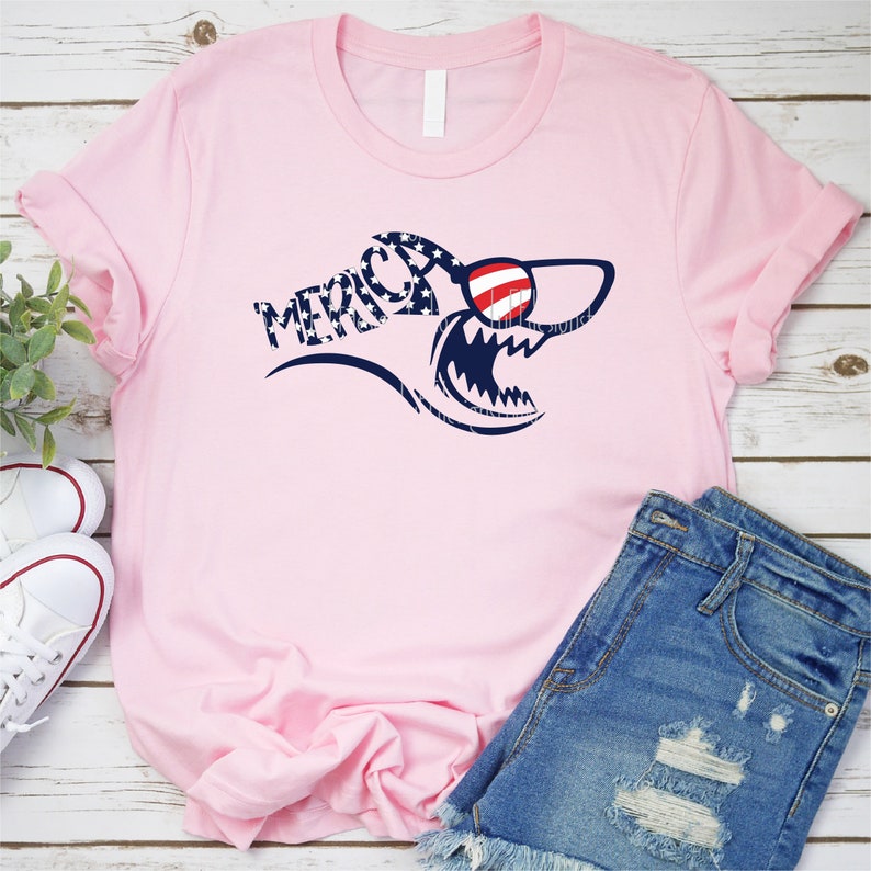 May include: Pink t-shirt with a shark graphic wearing sunglasses. The shark is made up of red, white, and blue stars and the text "Merica" is printed on the shirt.